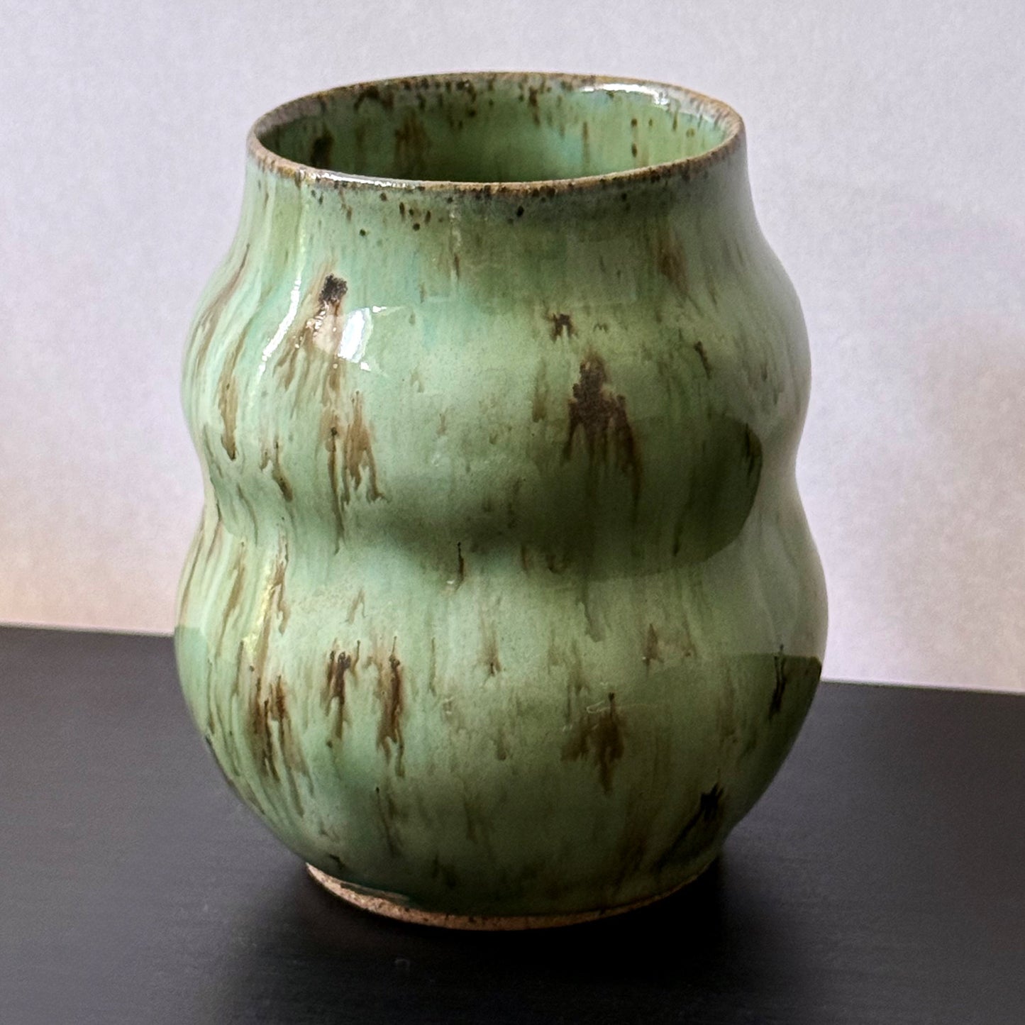 Handmade ceramic vase with a textured, ribbed body and a glossy light green glaze with brown distressed accents, sitting on a dark surface against a plain light gray wall.