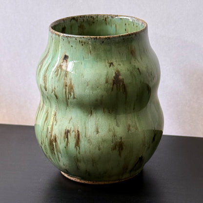 Handmade ceramic vase with a textured, ribbed body and a glossy light green glaze with brown distressed accents, sitting on a dark surface against a plain light gray wall.