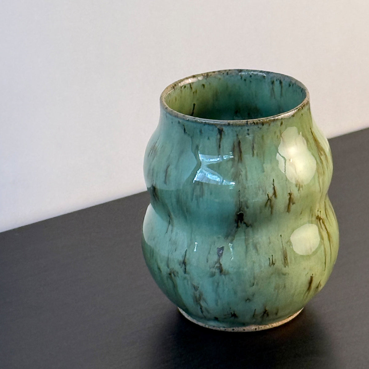 Handmade ceramic vase with a textured, ribbed body and a glossy light green glaze with brown distressed accents, sitting on a dark surface against a plain light gray wall.