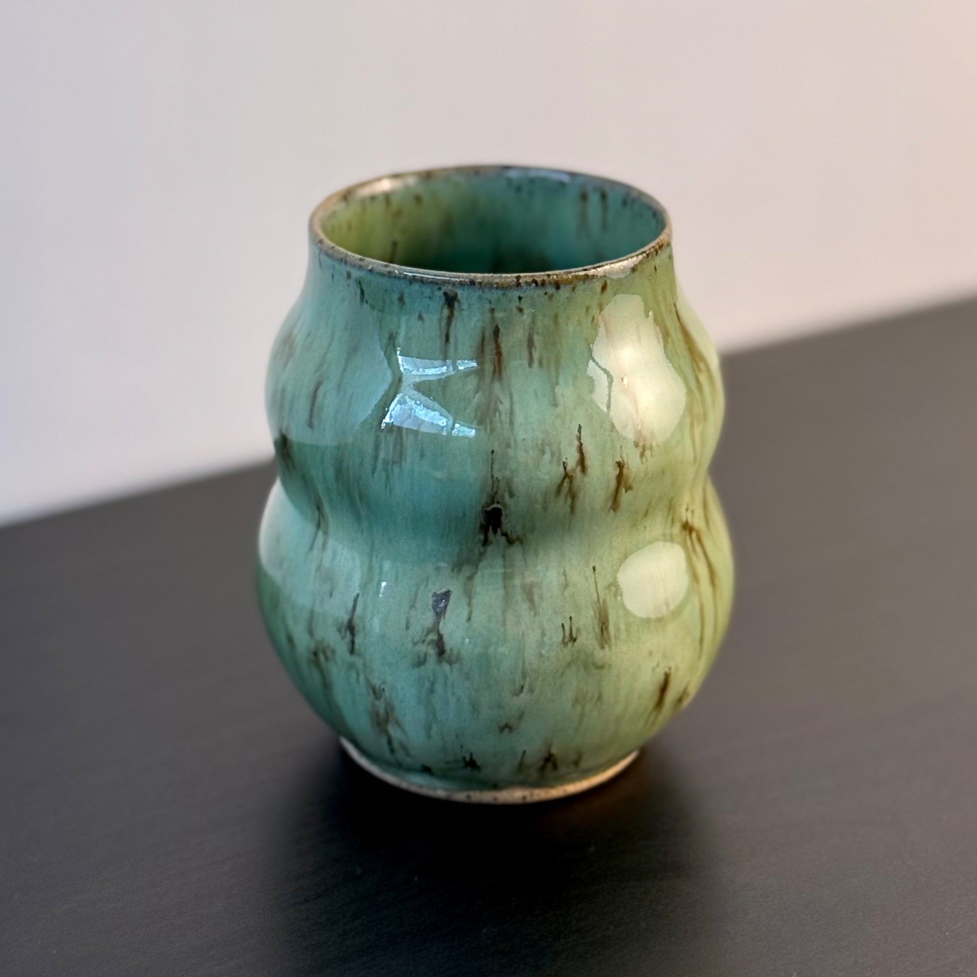 Handmade ceramic vase with a textured, ribbed body and a glossy light green glaze with brown distressed accents, sitting on a dark surface against a plain light gray wall.