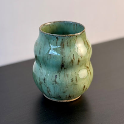 Handmade ceramic vase with a textured, ribbed body and a glossy light green glaze with brown distressed accents, sitting on a dark surface against a plain light gray wall.