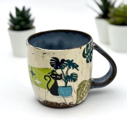 A handmade mug featuring a mischievous black cat hiding among illustrated monstera plants, with a deep blue interior and dark handle, shown with small succulents in the background.