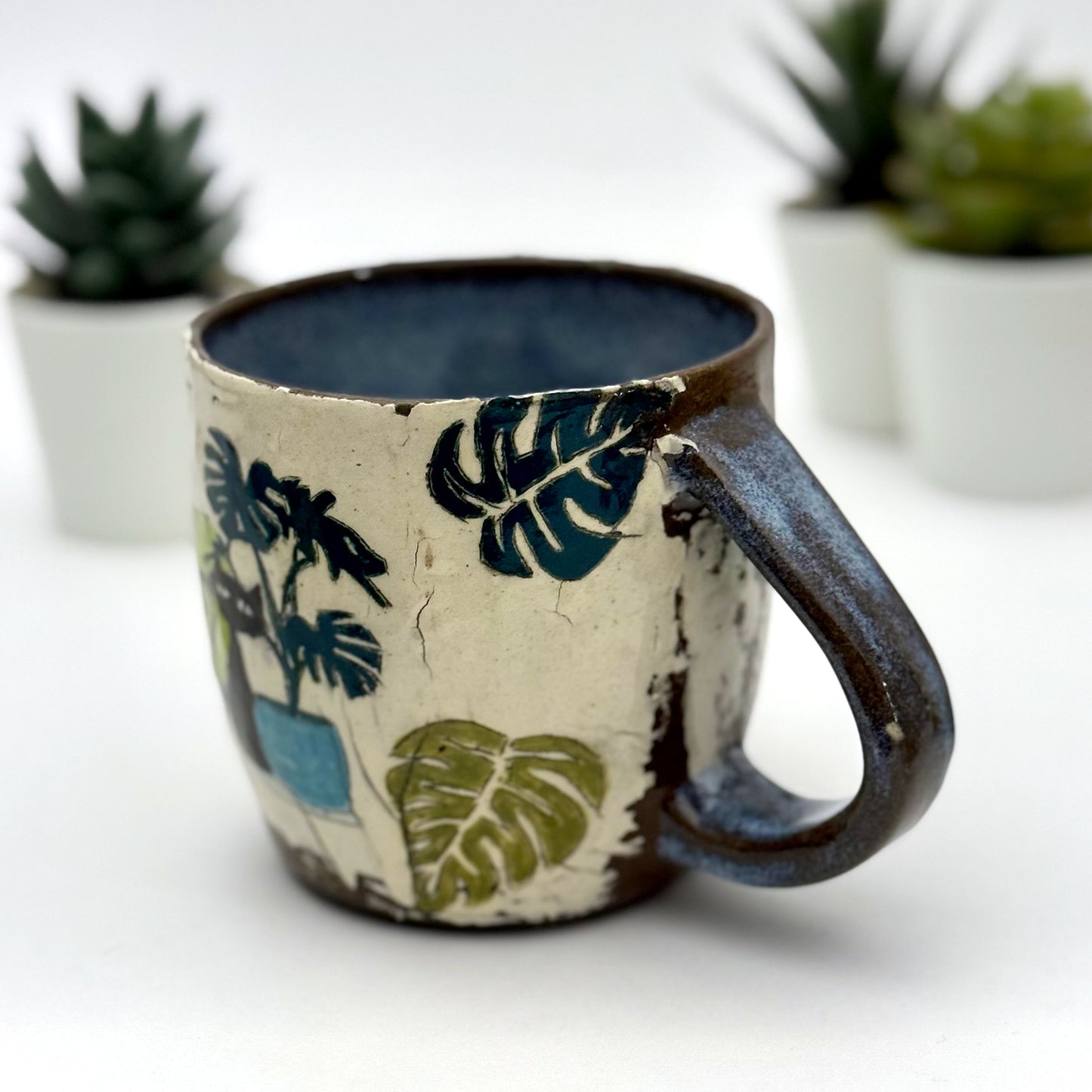 A handmade mug featuring a mischievous black cat hiding among illustrated monstera plants, with a deep blue interior and dark handle, shown with small succulents in the background.