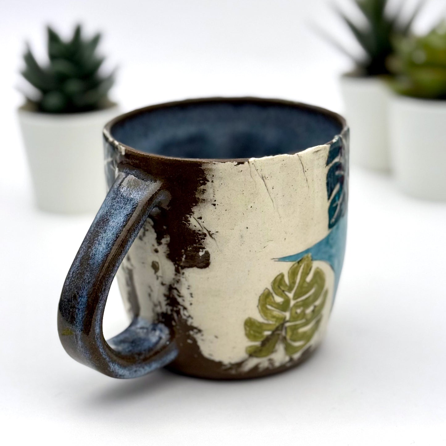 A handmade mug featuring a mischievous black cat hiding among illustrated monstera plants, with a deep blue interior and dark handle, shown with small succulents in the background.