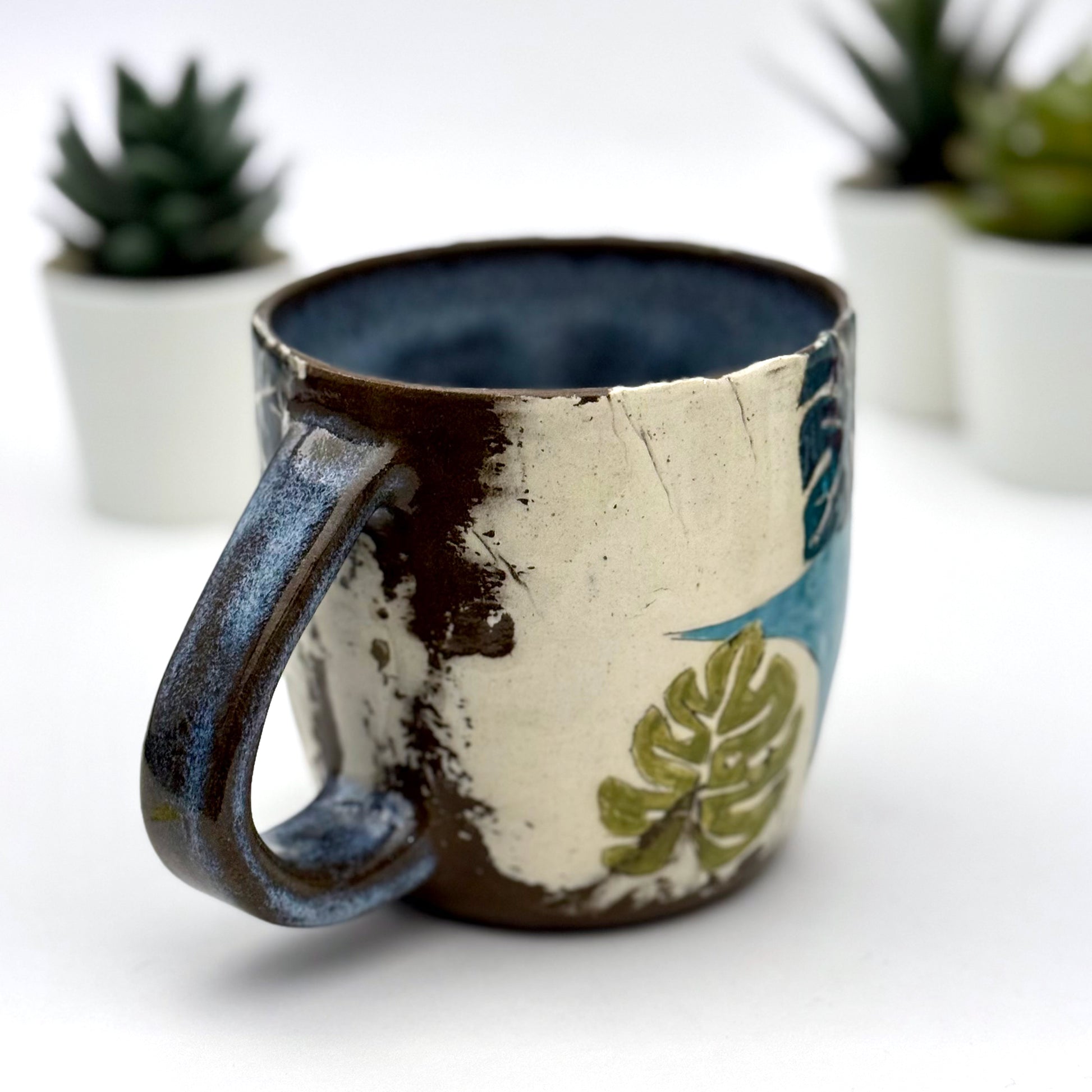 A handmade mug featuring a mischievous black cat hiding among illustrated monstera plants, with a deep blue interior and dark handle, shown with small succulents in the background.