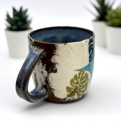 A handmade mug featuring a mischievous black cat hiding among illustrated monstera plants, with a deep blue interior and dark handle, shown with small succulents in the background.