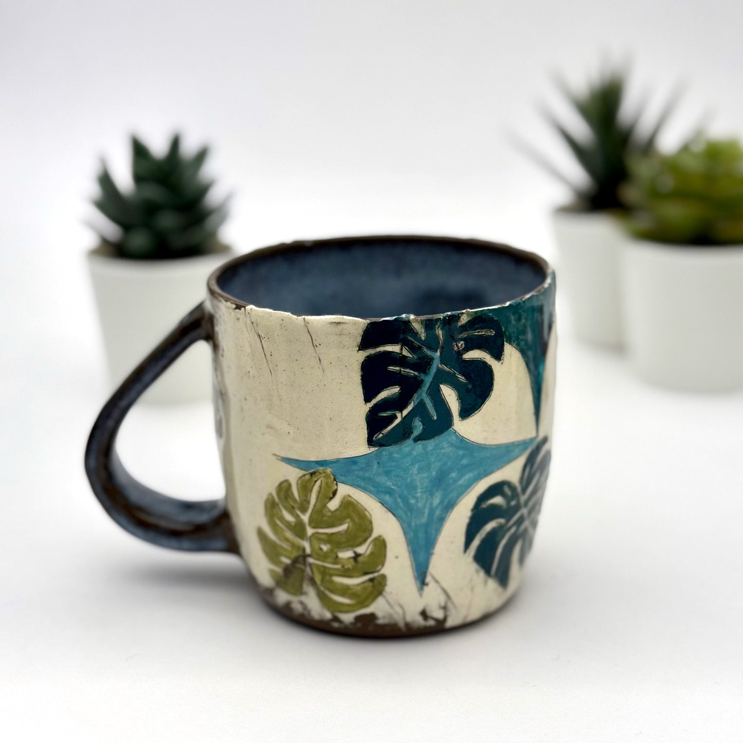 A handmade mug featuring a mischievous black cat hiding among illustrated monstera plants, with a deep blue interior and dark handle, shown with small succulents in the background.