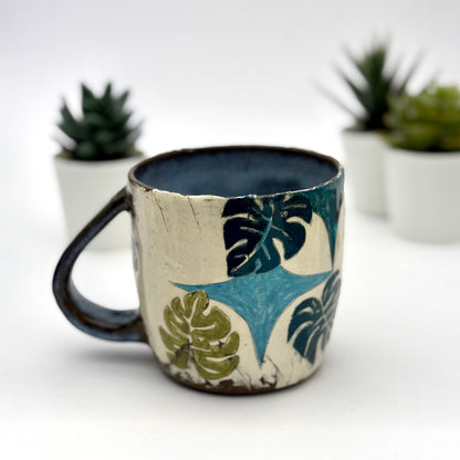 A handmade mug featuring a mischievous black cat hiding among illustrated monstera plants, with a deep blue interior and dark handle, shown with small succulents in the background.