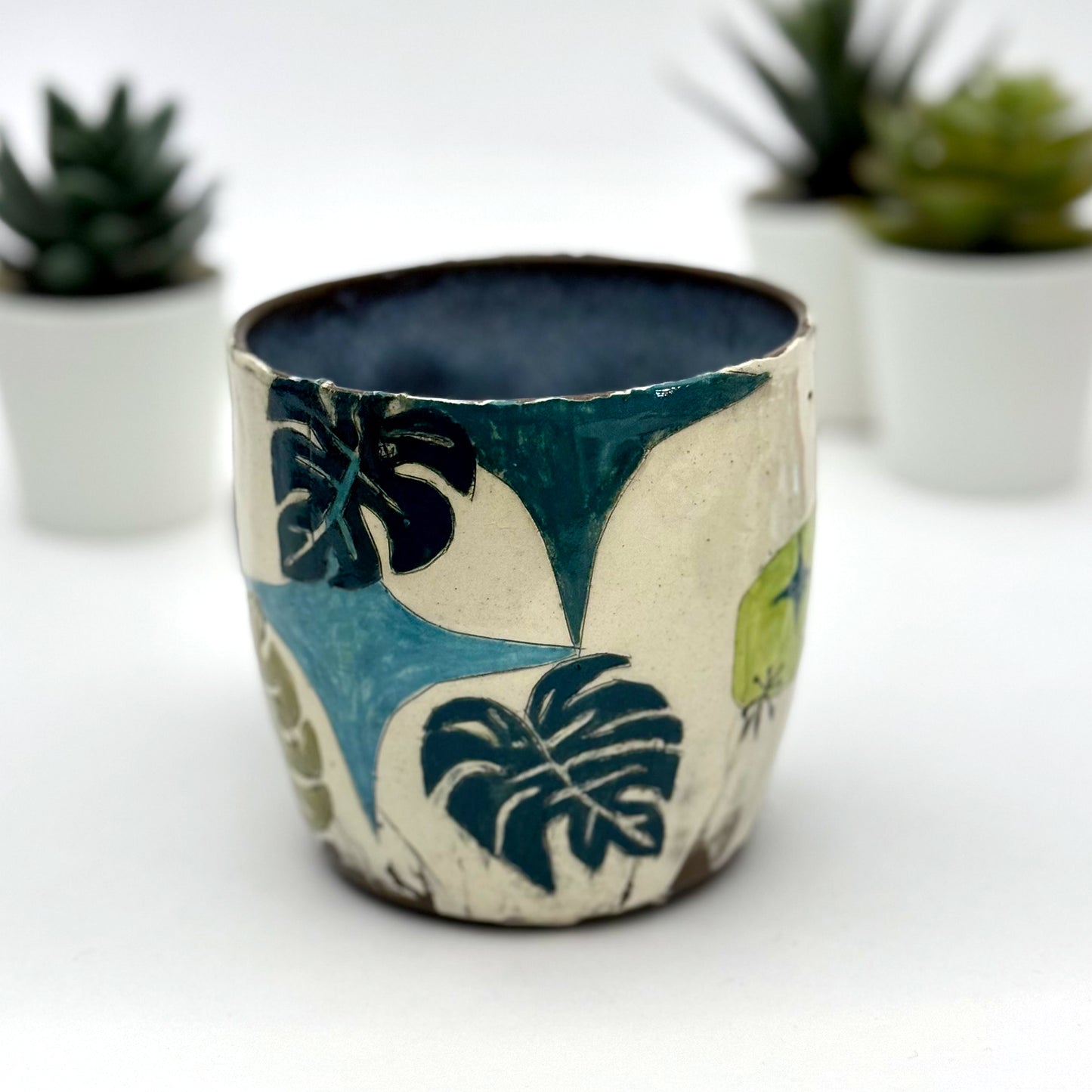 A handmade mug featuring a mischievous black cat hiding among illustrated monstera plants, with a deep blue interior and dark handle, shown with small succulents in the background.
