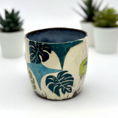 A handmade mug featuring a mischievous black cat hiding among illustrated monstera plants, with a deep blue interior and dark handle, shown with small succulents in the background.