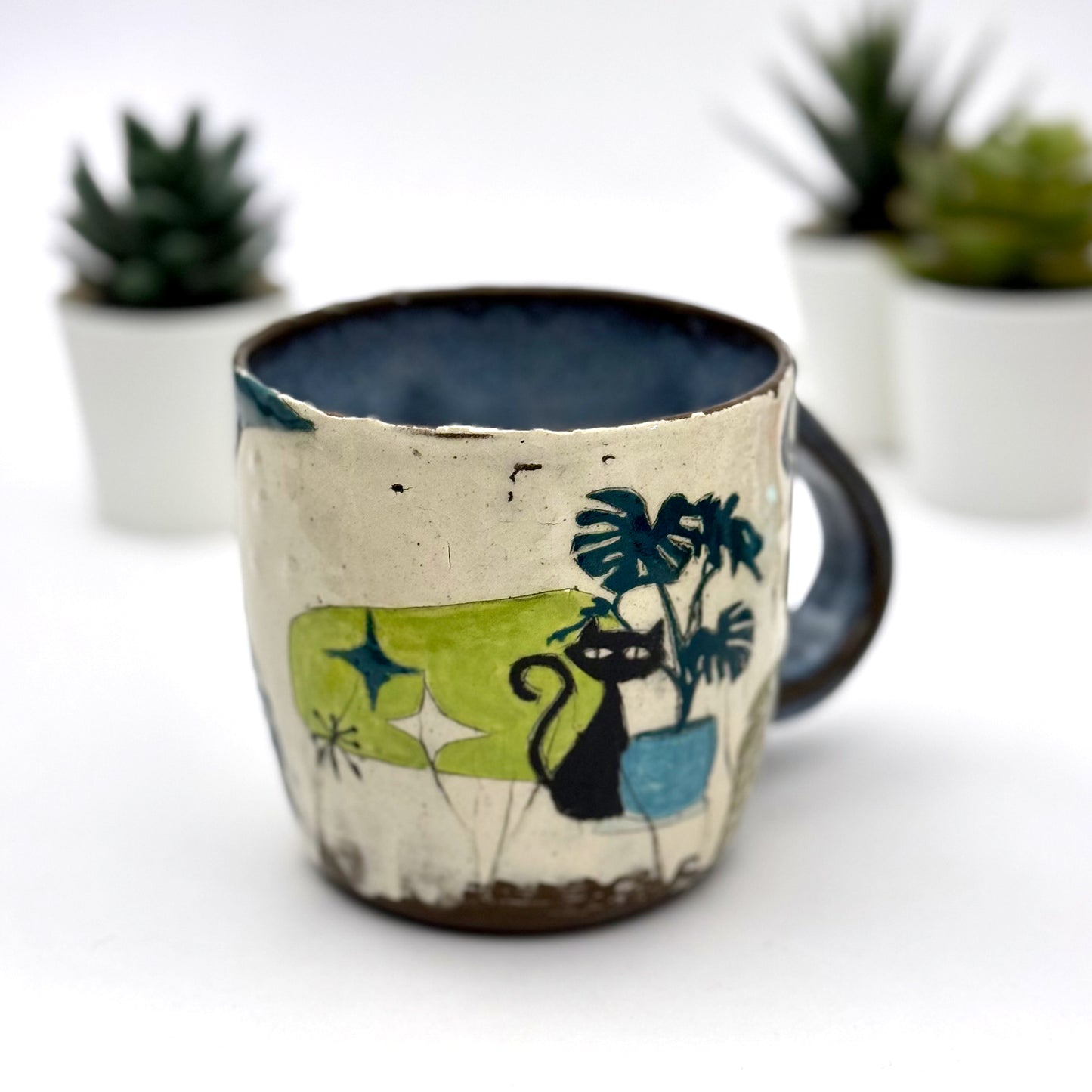 A handmade mug featuring a mischievous black cat hiding among illustrated monstera plants, with a deep blue interior and dark handle, shown with small succulents in the background.