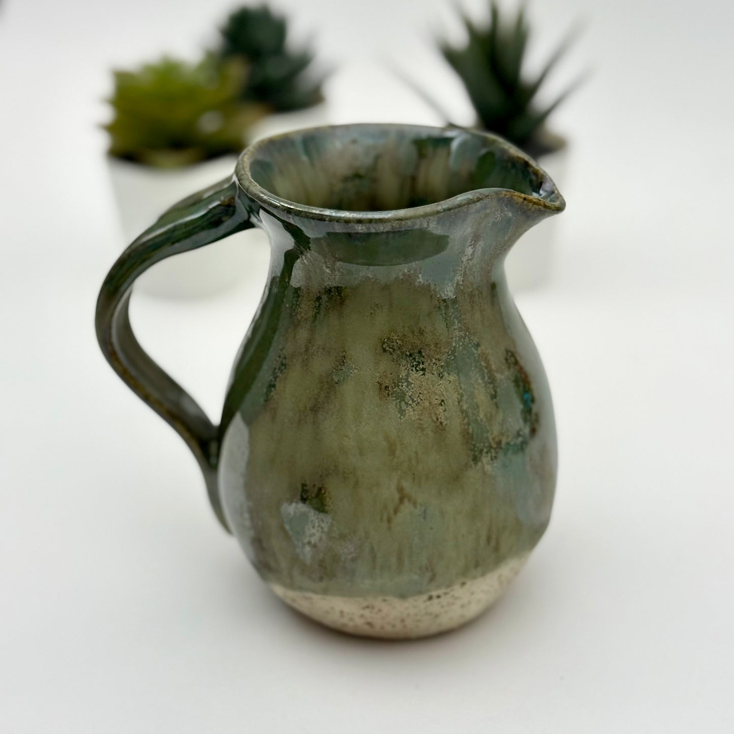 Green ceramic pitcher on a white background with blurred plants in the background