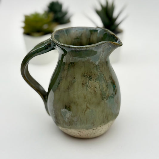 Green ceramic pitcher on a white background with blurred plants in the background