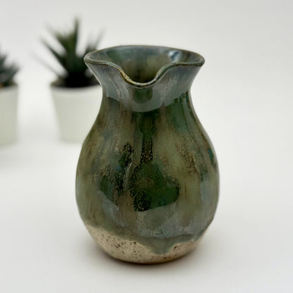 Green ceramic vase on a white surface with blurred plants in the background