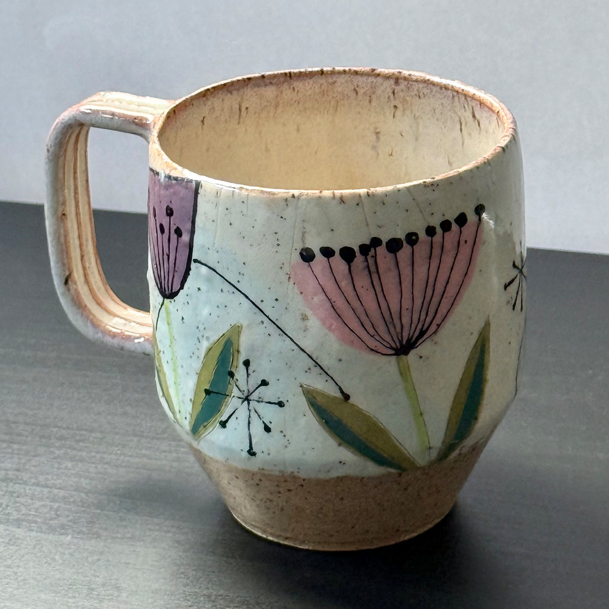 Handcrafted mid-century modern ceramic mug with pink and purple retro flower illustrations, blue seed pods, and atomic starbursts on a speckled cream background.