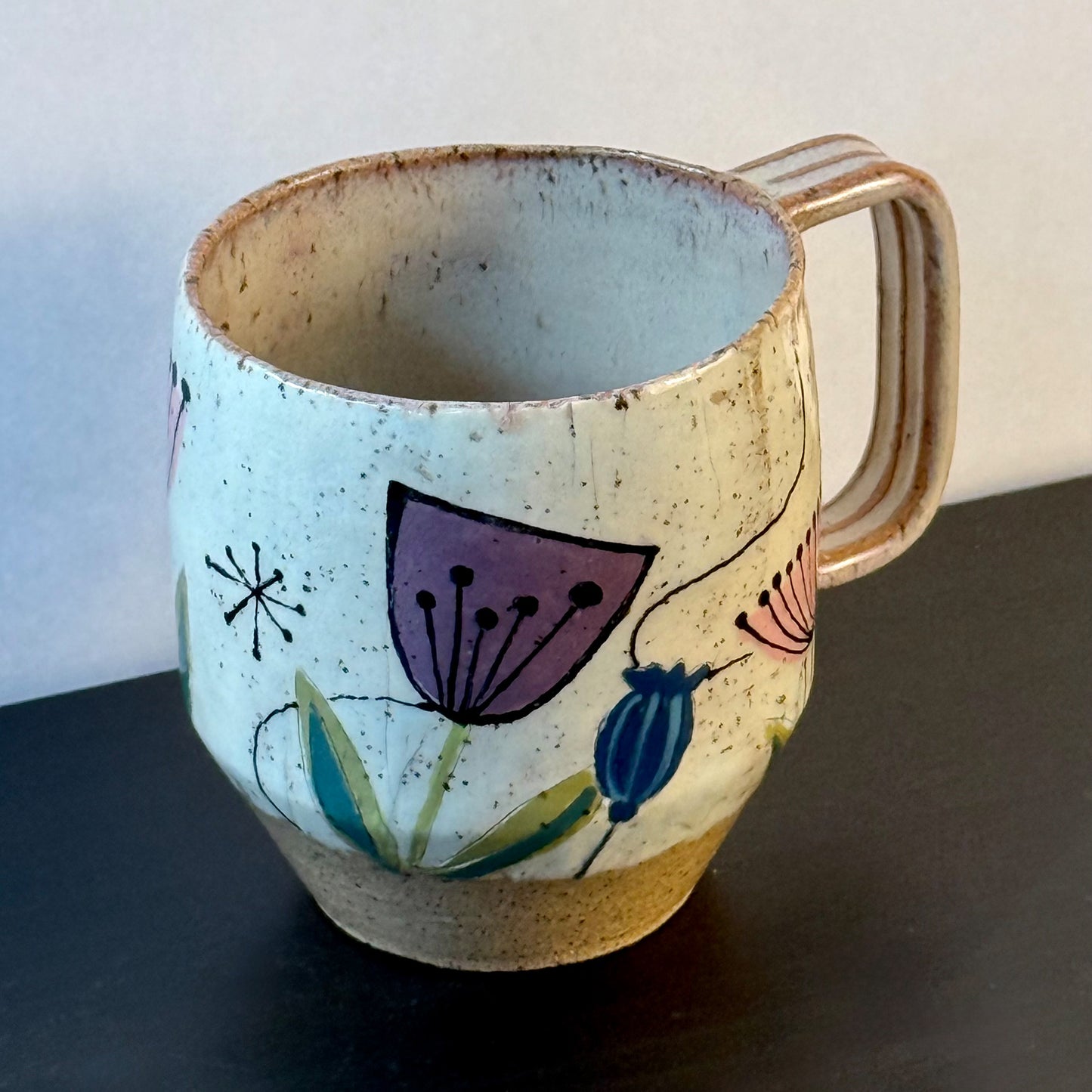 Handcrafted mid-century modern ceramic mug with pink and purple retro flower illustrations, blue seed pods, and atomic starbursts on a speckled cream background.