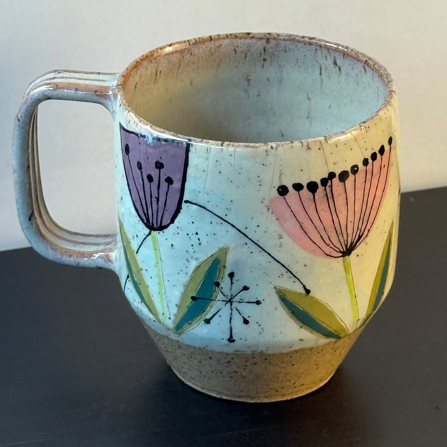 Handcrafted mid-century modern ceramic mug with pink and purple retro flower illustrations, blue seed pods, and atomic starbursts on a speckled cream background.
