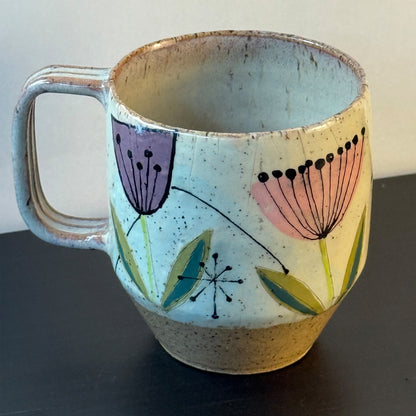 Handcrafted mid-century modern ceramic mug with pink and purple retro flower illustrations, blue seed pods, and atomic starbursts on a speckled cream background.