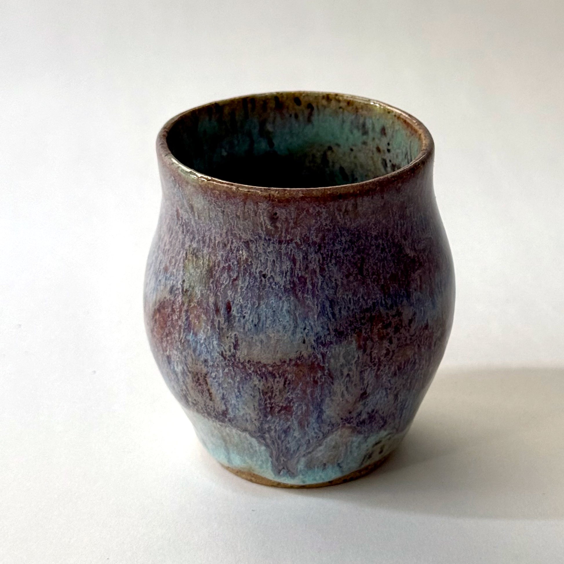Small handmade ceramic vase with a rounded silhouette and layered reactive glaze in shades of violet, plum, smoky blue, and pale turquoise. The surface has a textured, flowing appearance with organic glaze movement and a dark glossy interior rim. Photographed against a clean white background with soft natural lighting.