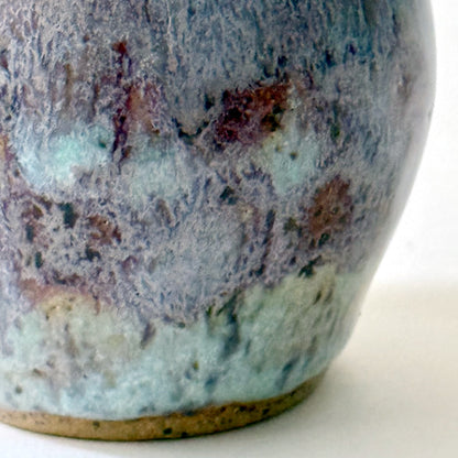Close up of small handmade ceramic vase with a rounded silhouette and layered reactive glaze in shades of violet, plum, smoky blue, and pale turquoise. The surface has a textured, flowing appearance with organic glaze movement and a dark glossy interior rim. Photographed against a clean white background with soft natural lighting.