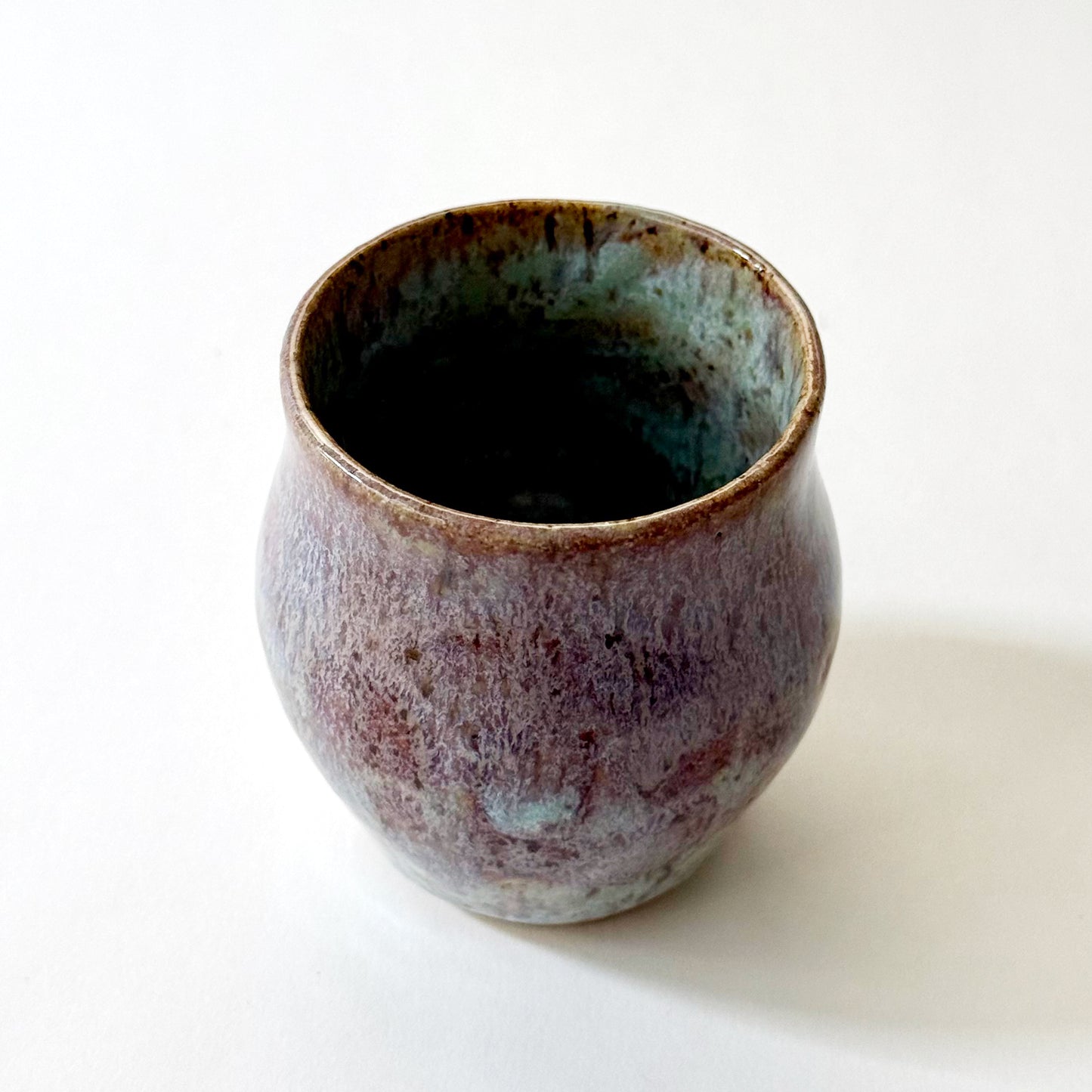 Small handmade ceramic vase with a rounded silhouette and layered reactive glaze in shades of violet, plum, smoky blue, and pale turquoise. The surface has a textured, flowing appearance with organic glaze movement and a dark glossy interior rim. Photographed against a clean white background with soft natural lighting.