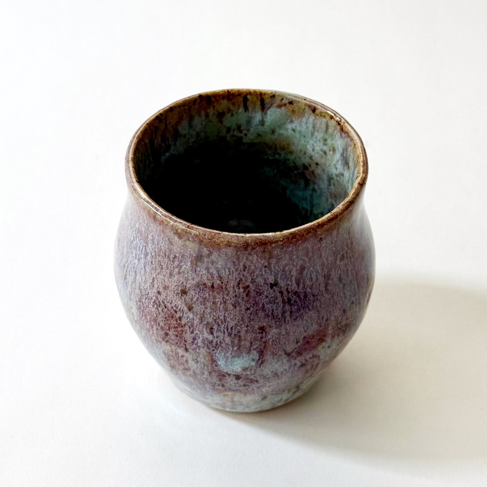 Small handmade ceramic vase with a rounded silhouette and layered reactive glaze in shades of violet, plum, smoky blue, and pale turquoise. The surface has a textured, flowing appearance with organic glaze movement and a dark glossy interior rim. Photographed against a clean white background with soft natural lighting.
