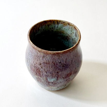 Small handmade ceramic vase with a rounded silhouette and layered reactive glaze in shades of violet, plum, smoky blue, and pale turquoise. The surface has a textured, flowing appearance with organic glaze movement and a dark glossy interior rim. Photographed against a clean white background with soft natural lighting.