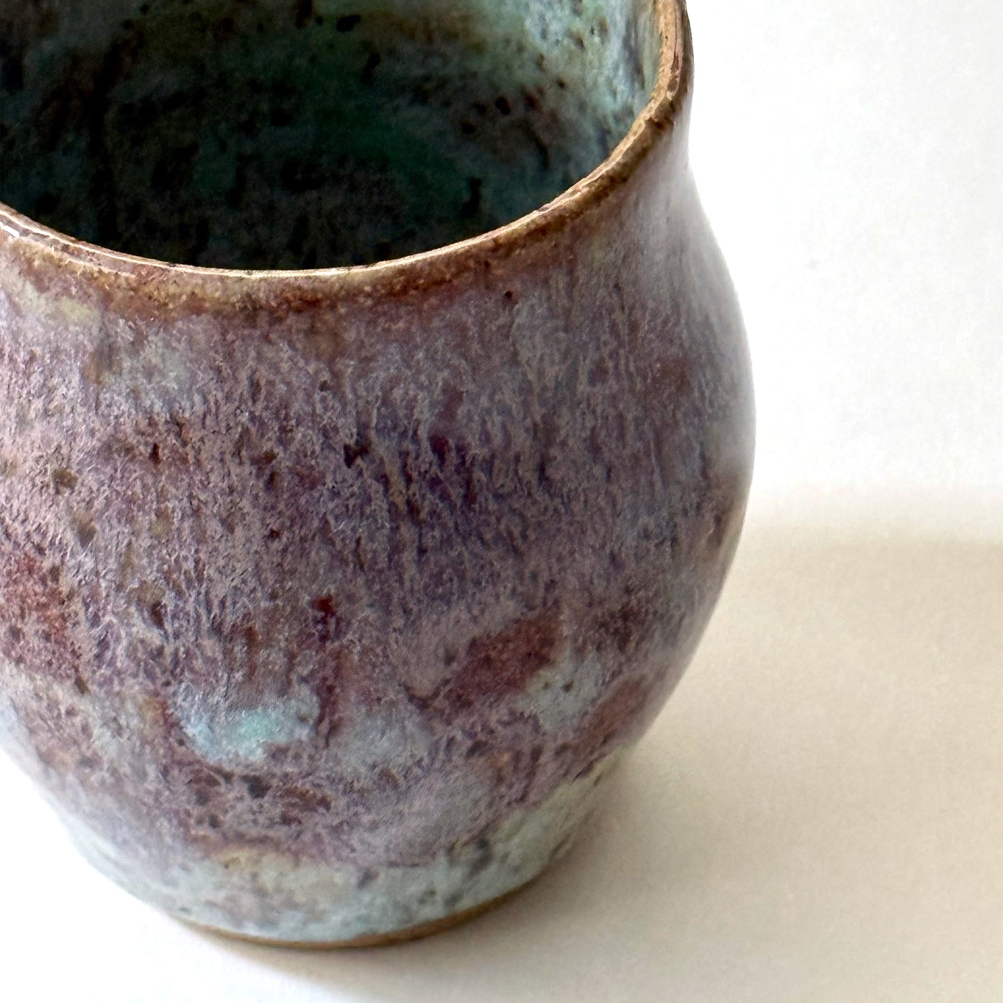Close up of small handmade ceramic vase with a rounded silhouette and layered reactive glaze in shades of violet, plum, smoky blue, and pale turquoise. The surface has a textured, flowing appearance with organic glaze movement and a dark glossy interior rim. Photographed against a clean white background with soft natural lighting.