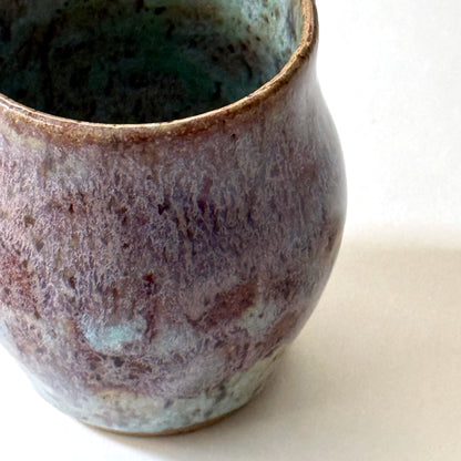 Close up of small handmade ceramic vase with a rounded silhouette and layered reactive glaze in shades of violet, plum, smoky blue, and pale turquoise. The surface has a textured, flowing appearance with organic glaze movement and a dark glossy interior rim. Photographed against a clean white background with soft natural lighting.