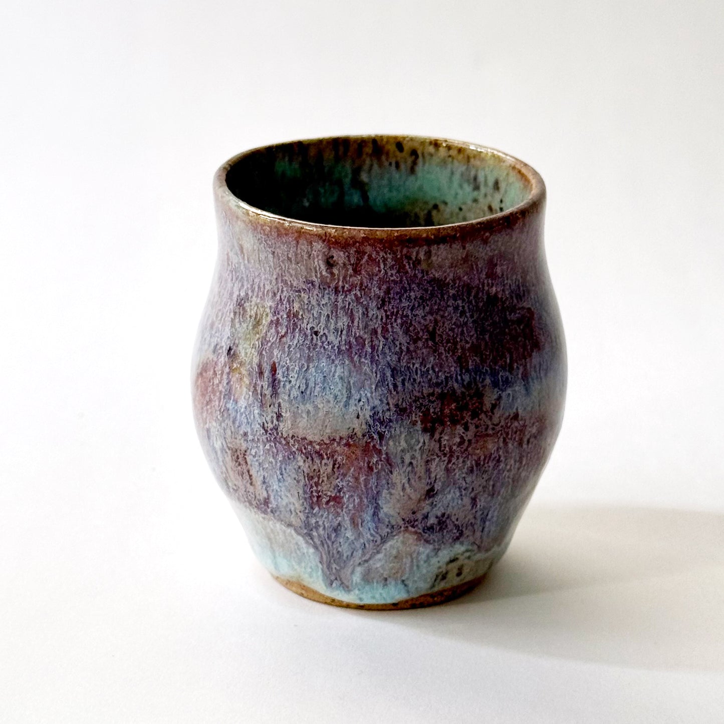 Small handmade ceramic vase with a rounded silhouette and layered reactive glaze in shades of violet, plum, smoky blue, and pale turquoise. The surface has a textured, flowing appearance with organic glaze movement and a dark glossy interior rim. Photographed against a clean white background with soft natural lighting.