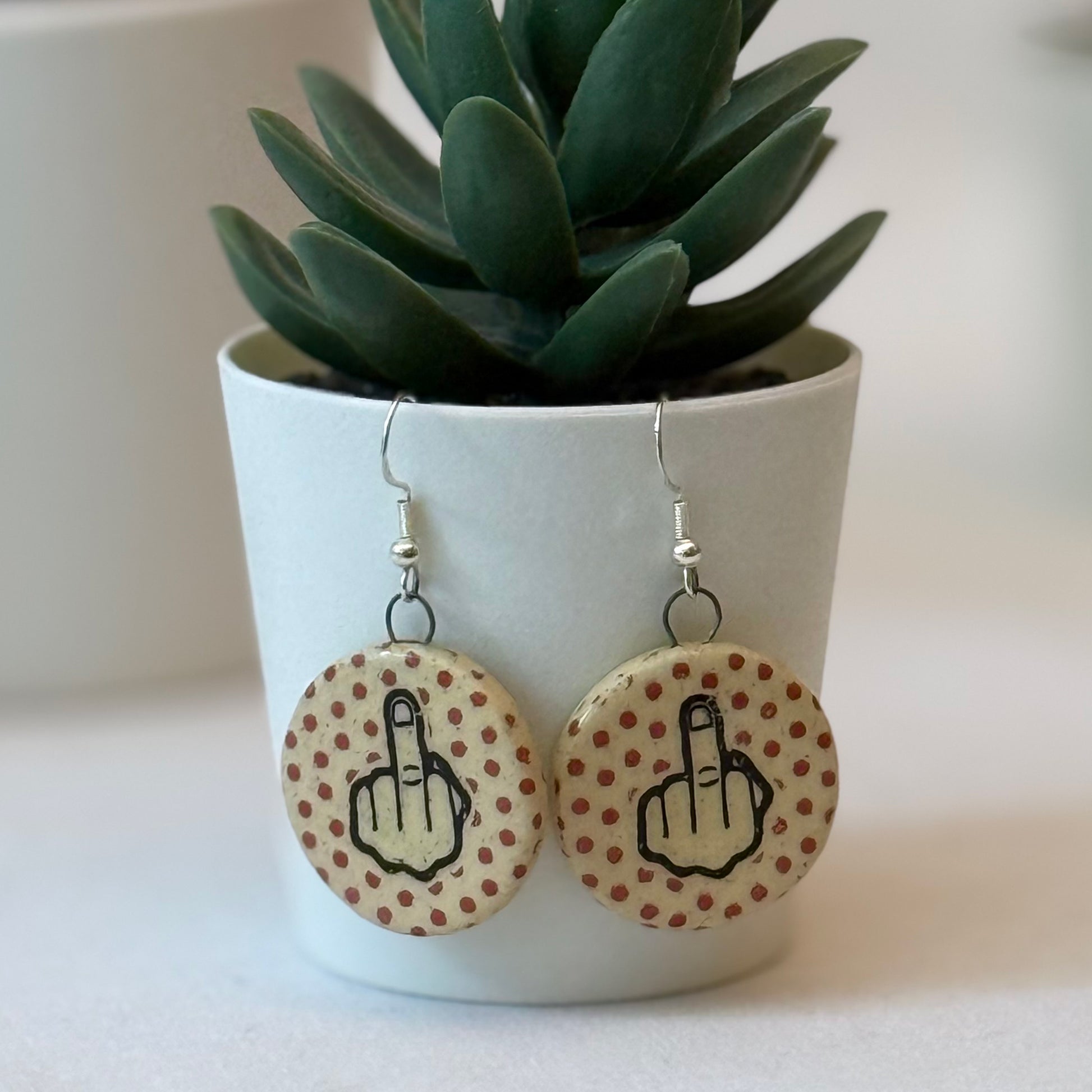 Earrings with middle finger design on a polka dot background, placed in front of a potted plant.