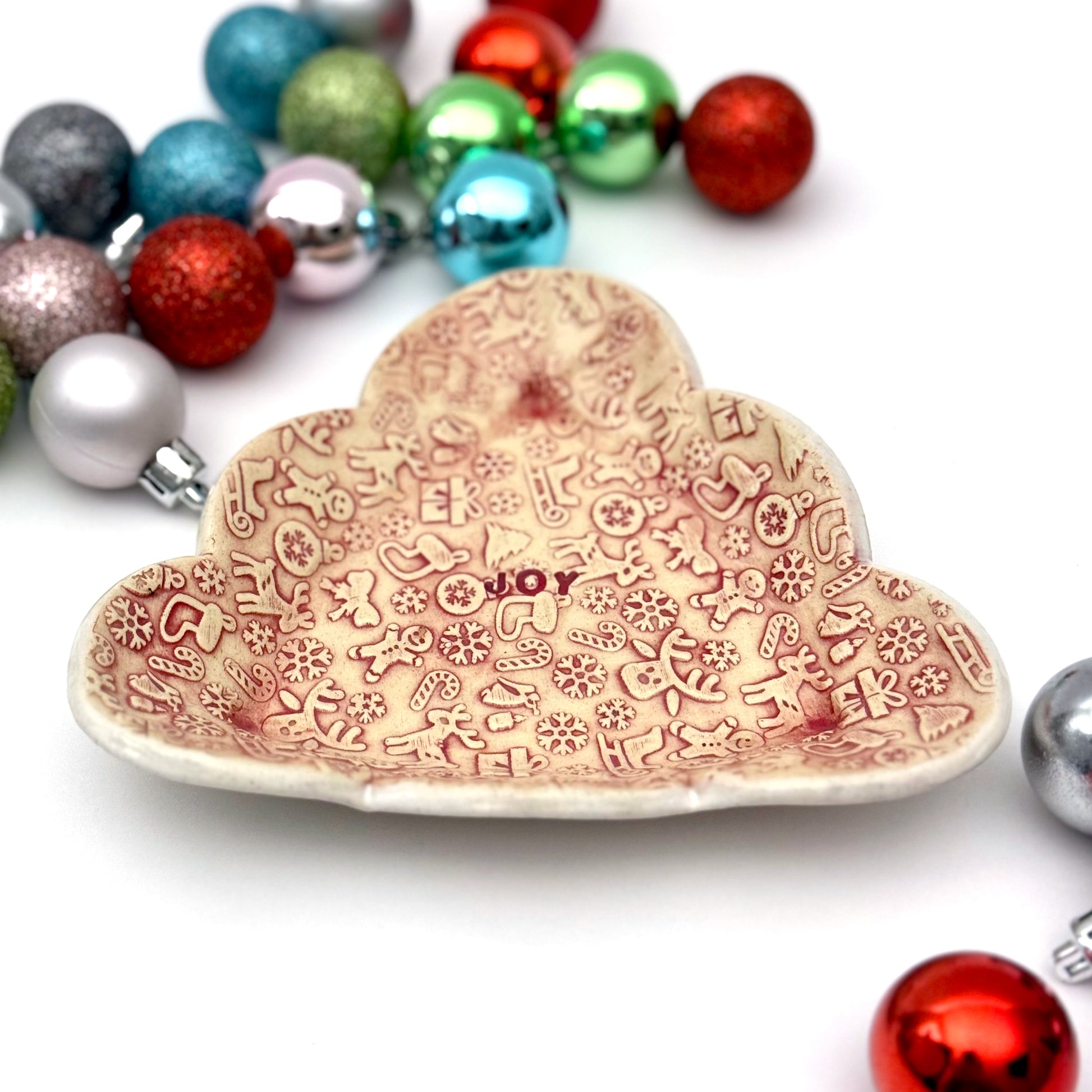 Red and cream covered ceramic plate with tiny embossed holiday designs and the word “JOY” in the middle, styled with assorted ornaments around it.