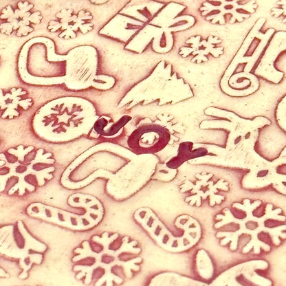 Close-up of red and cream covered ceramic plate with tiny embossed holiday designs and the word “JOY” in the middle, styled with assorted ornaments around it.