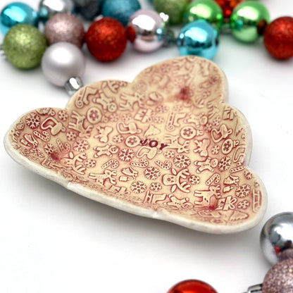 Red and cream covered ceramic plate with tiny embossed holiday designs and the word “JOY” in the middle, styled with assorted ornaments around it.
