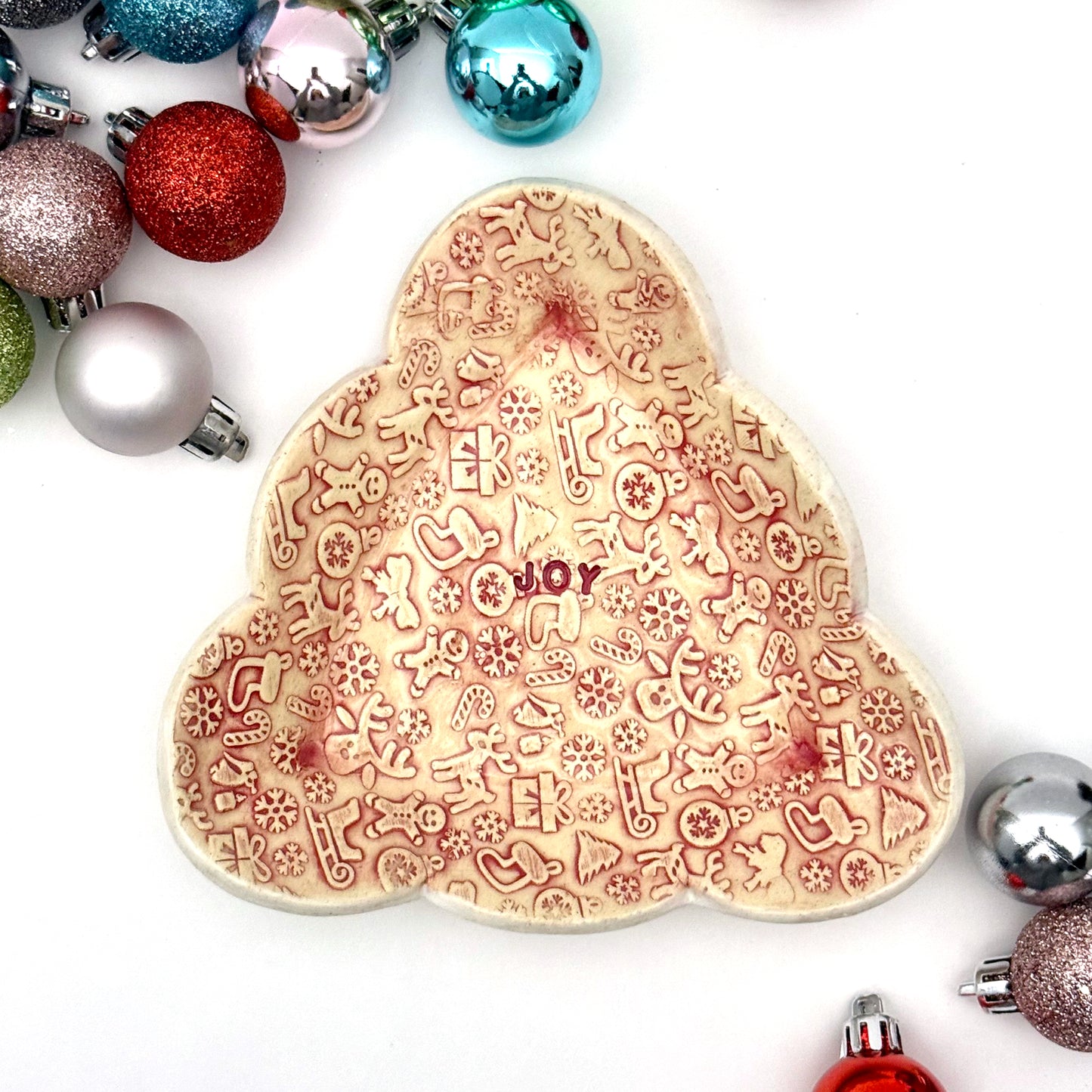 Red and cream covered ceramic plate with tiny embossed holiday designs and the word “JOY” in the middle, styled with assorted ornaments around it.