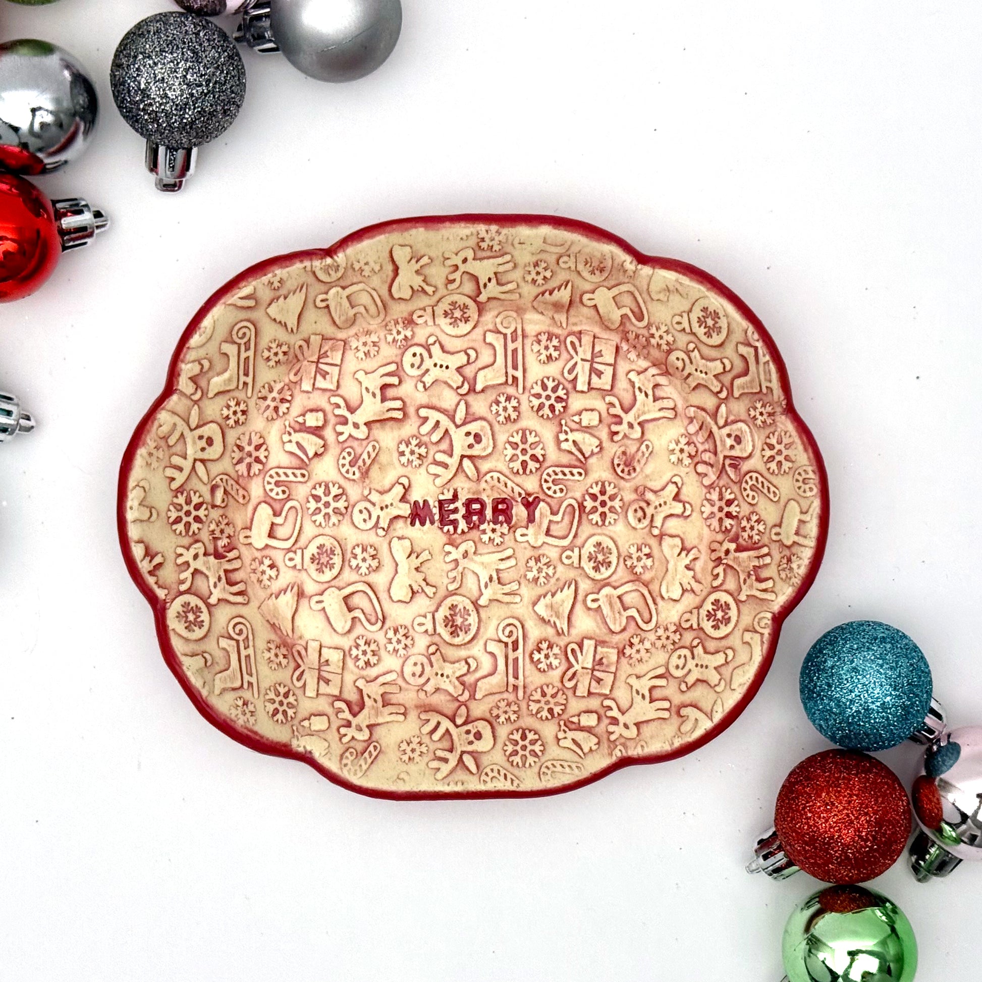 A scalloped ceramic dish with a red rim and embossed holiday icons—reindeer, gingerbread people, trees, gifts, and snowflakes—with the word “MERRY” in the center. Surrounding the dish are small colorful ornaments.
