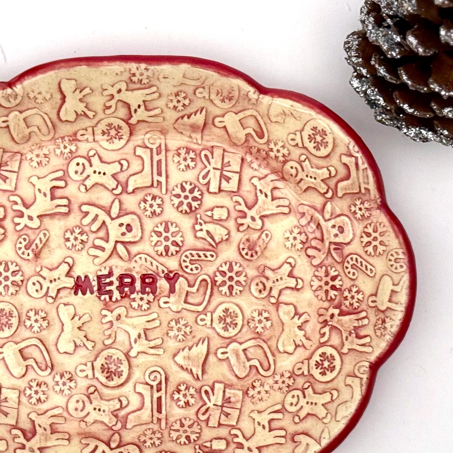 Close-up of a scalloped ceramic dish with a red rim and embossed holiday icons—reindeer, gingerbread people, trees, gifts, and snowflakes—with the word “MERRY” in the center. 