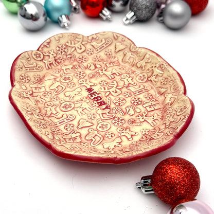 A scalloped ceramic dish with a red rim and embossed holiday icons—reindeer, gingerbread people, trees, gifts, and snowflakes—with the word “MERRY” in the center. Surrounding the dish are small colorful ornaments.