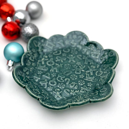 A scalloped ceramic dish glazed in deep evergreen with an embossed holiday pattern featuring reindeer, gingerbread people, snowflakes, stockings, and candy canes. Colorful ornaments sit to the side for decoration.