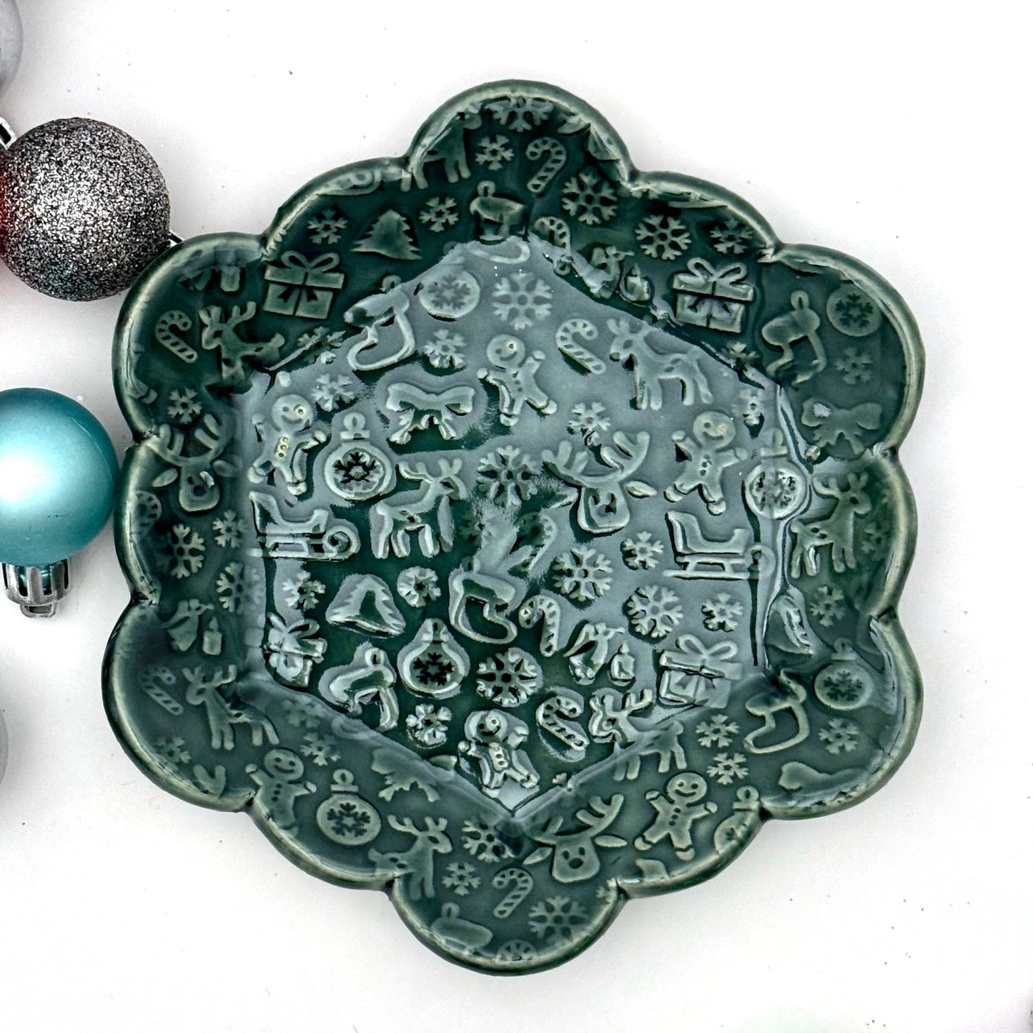 A scalloped ceramic dish glazed in deep evergreen with an embossed holiday pattern featuring reindeer, gingerbread people, snowflakes, stockings, and candy canes. Colorful ornaments sit to the side for decoration.