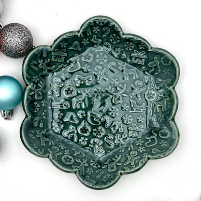 A scalloped ceramic dish glazed in deep evergreen with an embossed holiday pattern featuring reindeer, gingerbread people, snowflakes, stockings, and candy canes. Colorful ornaments sit to the side for decoration.