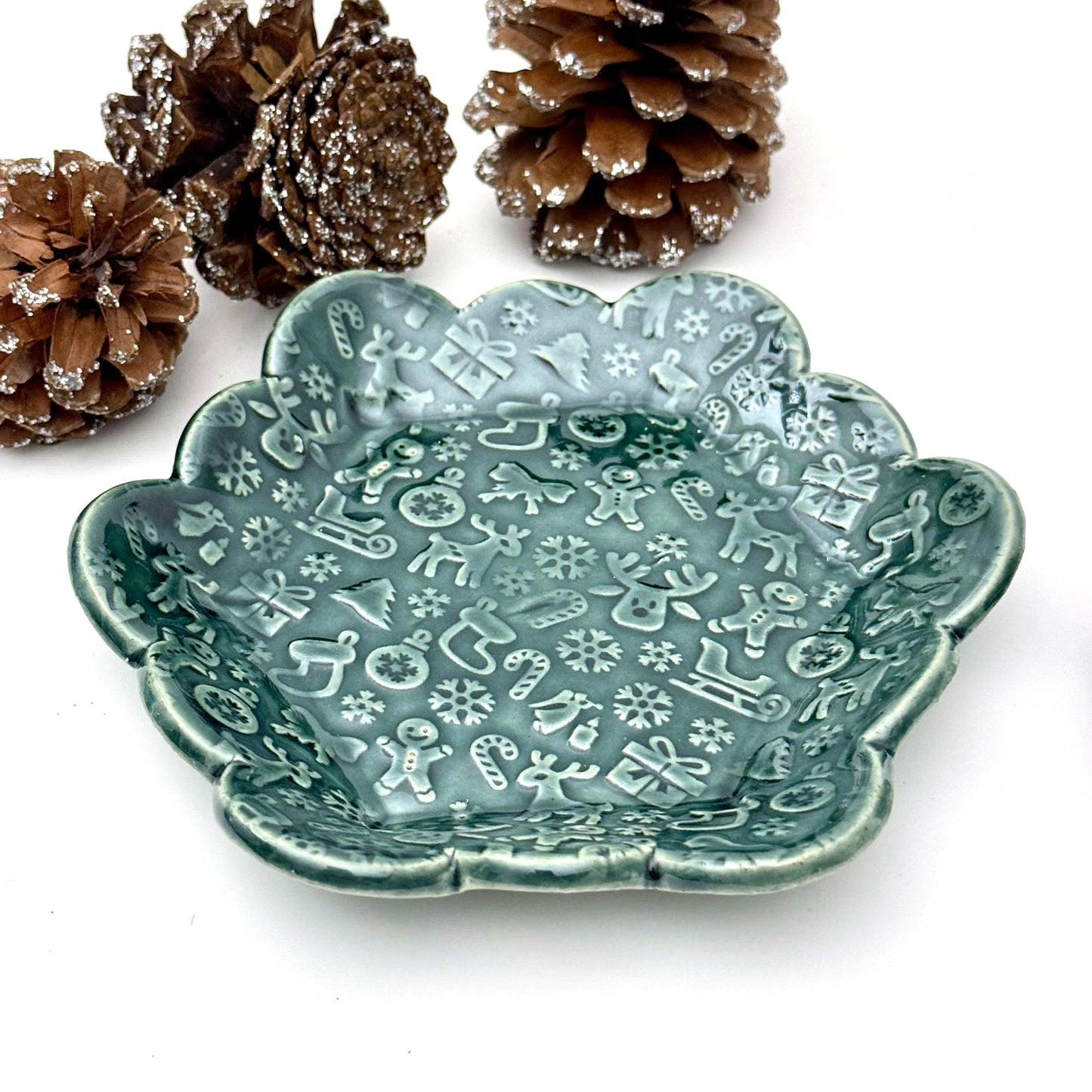 A scalloped ceramic dish glazed in deep evergreen with an embossed holiday pattern featuring reindeer, gingerbread people, snowflakes, stockings, and candy canes. 