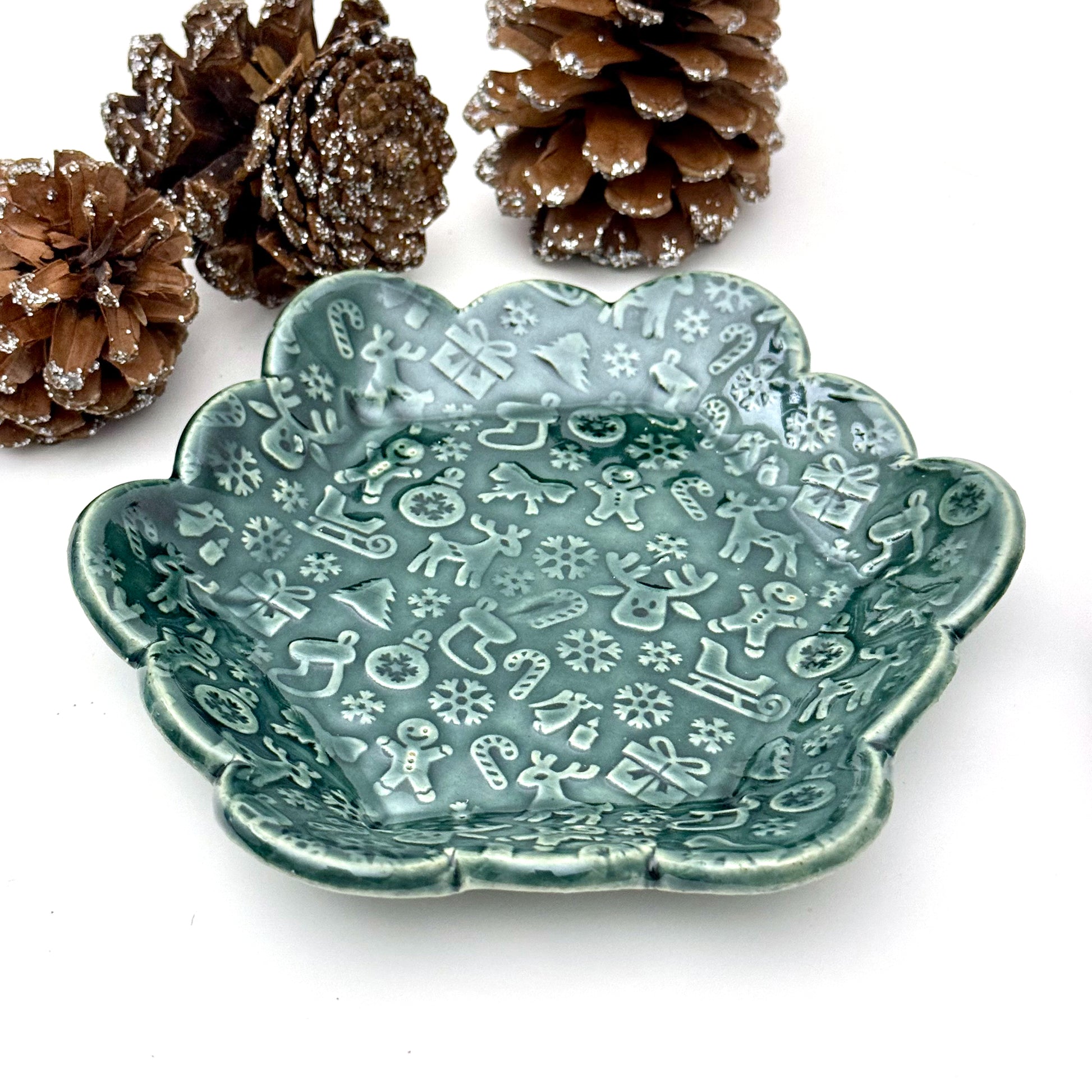 A scalloped ceramic dish glazed in deep evergreen with an embossed holiday pattern featuring reindeer, gingerbread people, snowflakes, stockings, and candy canes. 