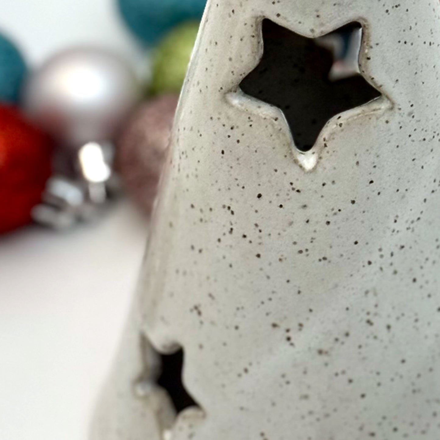 Close-up of a white ceramic tree luminary with speckled glaze and star-shaped cutouts, surrounded by colorful holiday ornaments.