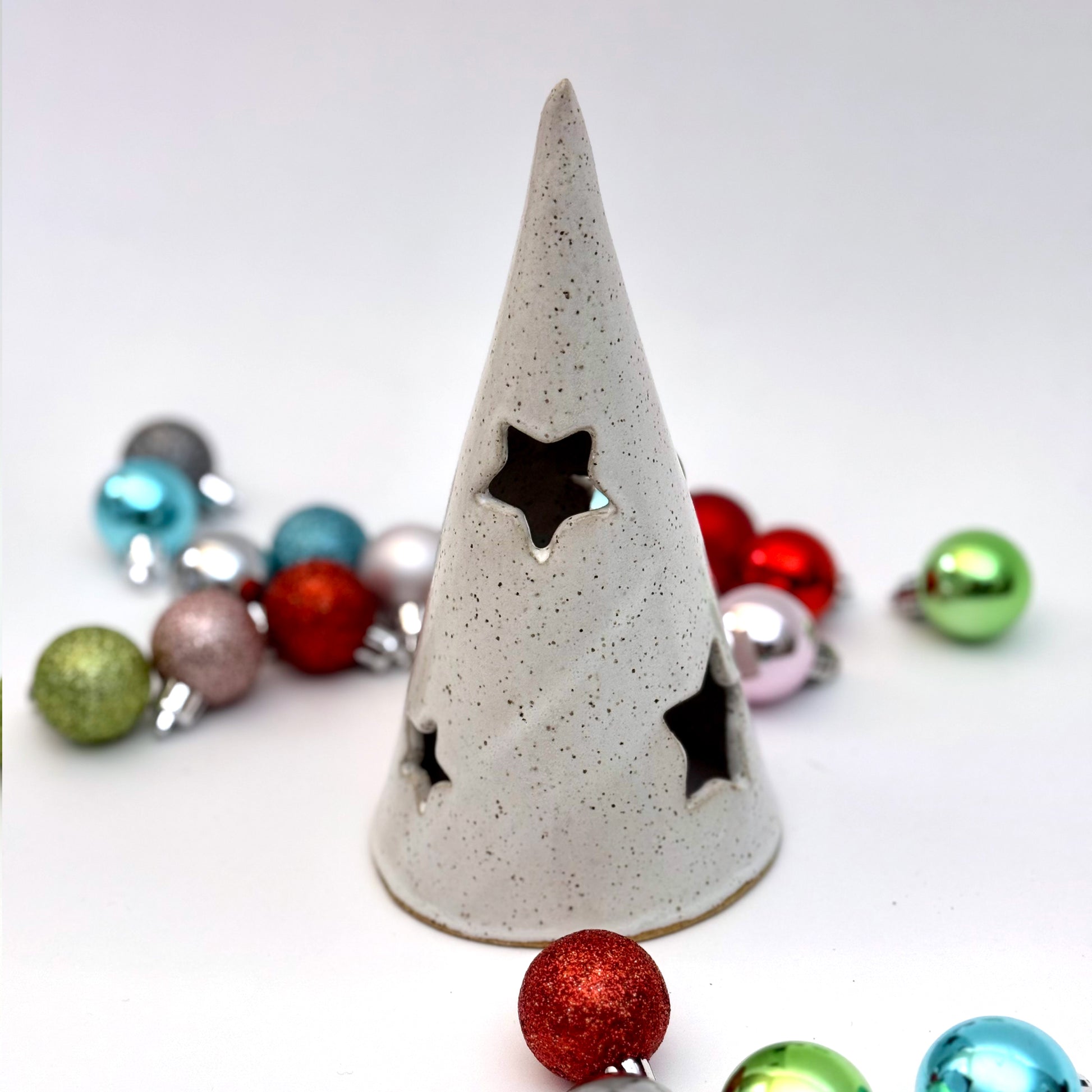A white ceramic tree luminary with speckled glaze and star-shaped cutouts, surrounded by colorful holiday ornaments.