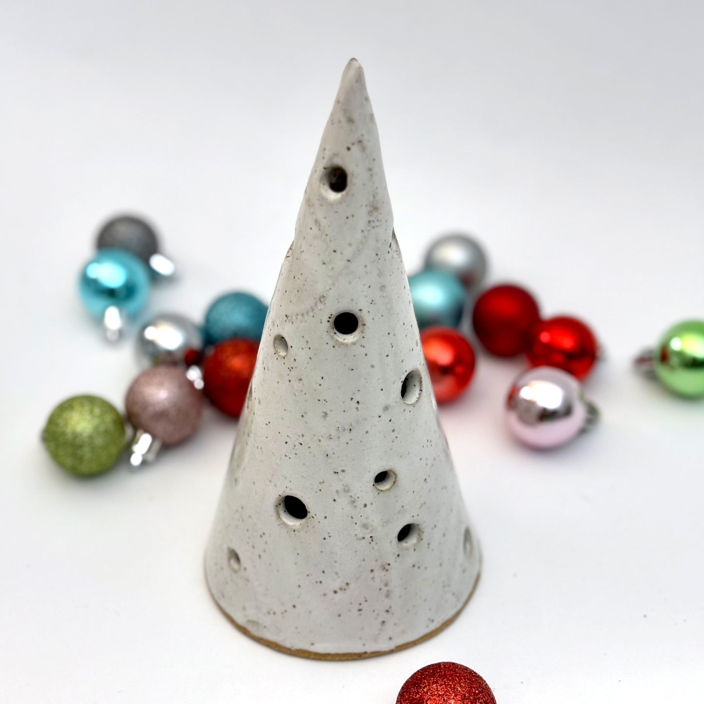 A white speckled ceramic tree luminary with round cutout holes, surrounded by colorful holiday ornaments.