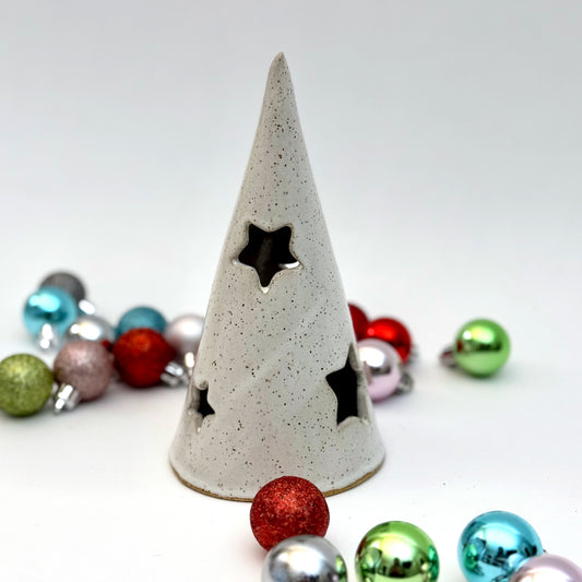A white ceramic tree luminary with speckled glaze and star-shaped cutouts, surrounded by colorful holiday ornaments.