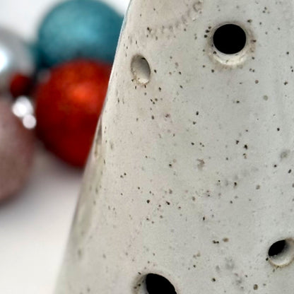 Close-up of A white speckled ceramic tree luminary with round cutout holes, surrounded by colorful holiday ornaments.