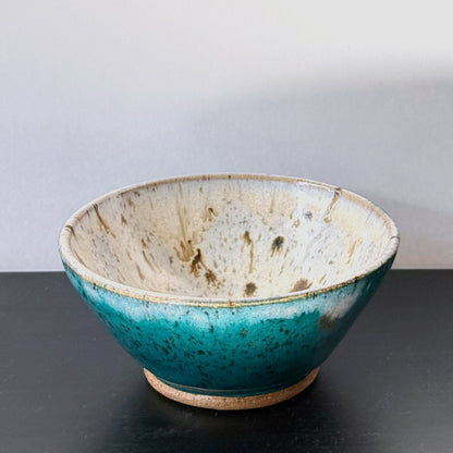 Handmade ceramic bowl with a saturated teal exterior and a creamy speckled interior, finished with a wide rounded profile and glossy glaze.