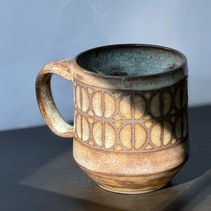Handmade ceramic mug with a repeating Split Circle breeze block pattern, dark clay showing through the design, and a warm earthy glaze.